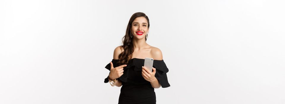 Online shopping concept. Stylish woman in black dress, wearing makeup, pointing finger at mobile phone with satisfied smile, standing over white background.
