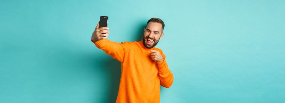 Handsome man video calling on mobile phone, taking selfie and pointing at smartphone camera, standing over turquoise background.