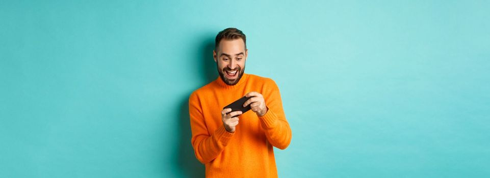 Excited adult man playing on mobile phone, looking amazed at smartphone screen, standing over turquoise background.