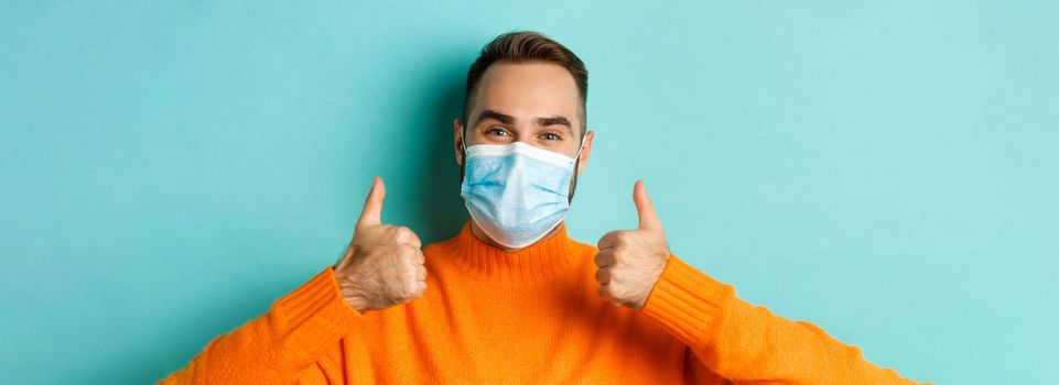 Covid-19, social distancing and quarantine concept. Close-up of satisfied male model in medical mask showing thumbs-up, praising and agreeing, standing over light blue background.