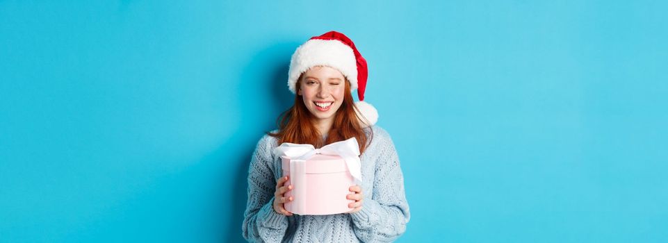 Winter holidays and Christmas Eve concept. Cute redhead girl in sweater and Santa hat, holding New Year gift and looking at camera, standing against blue background.