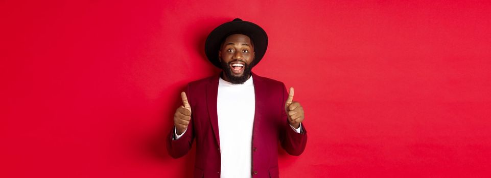 Christmas shopping and people concept. Handsome Black man smiling satisfied, showing thumbs-up, like and agree, approve something, standing against red background.
