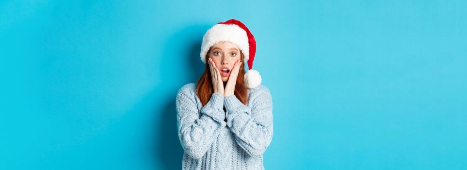 Winter holidays and Christmas Eve concept. Surprised redhead girl in santa hat, staring with disbelief at camera, open mouth amazed, standing over blue background.