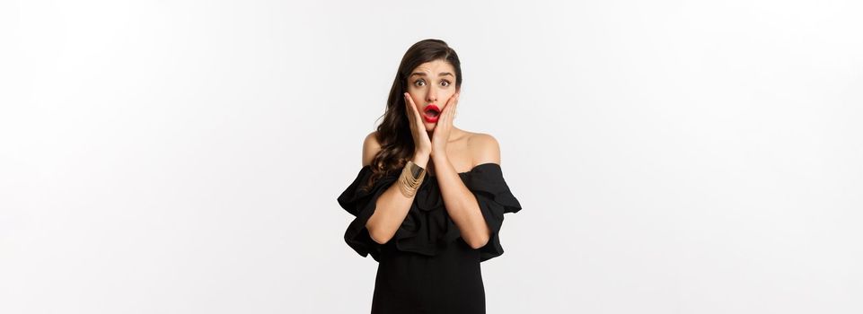 Beauty and fashion concept. Waist-up shot of surprised woman in black cocktail dress, looking amazed at camera, open mouth fascinated, standing over white background.