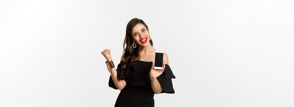 Online shopping concept. Satisfied pretty woman showing smartphone screen, making fist pump to rejoice, winning in internet, standing over white background.