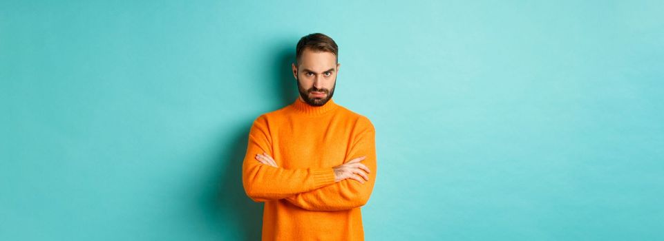 Offended man looking angry at you, cross arms on chest and stare mad, standing in orange sweater against turquoise background.