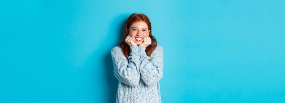 Lovely redhead teenage girl looking daydreaming, staring with admirationa sympathy at camera, standing against blue background.