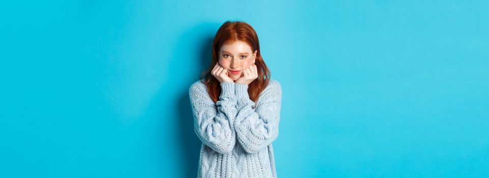 Lovely redhead teenage girl looking daydreaming, staring with admirationa sympathy at camera, standing against blue background.