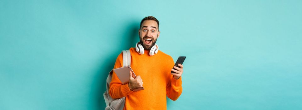 Handsome man student with headphones and backpack, holding digital tablet and smartphone, looking amazed at camera, standing against turquoise background.