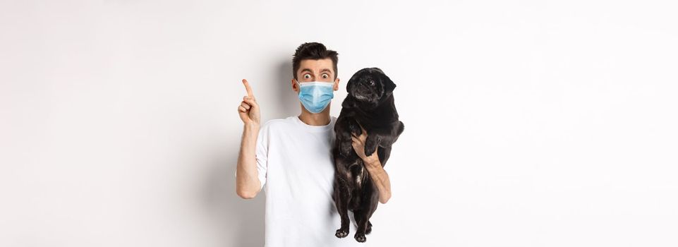 Covid-19, animals and quarantine concept. Young man in medical mask holding cute black pug, dog looking right and owner pointing at logo, white background.