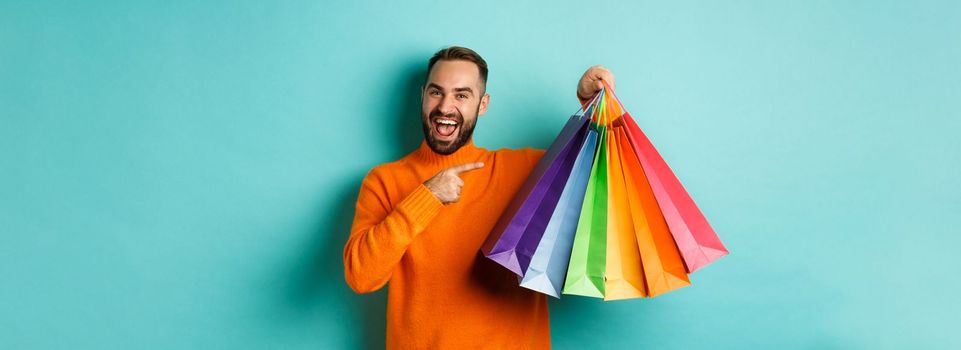 Happy handsome man holding shopping bags and smiling, pointing at purchased items and recommending store, standing over turquoise background.