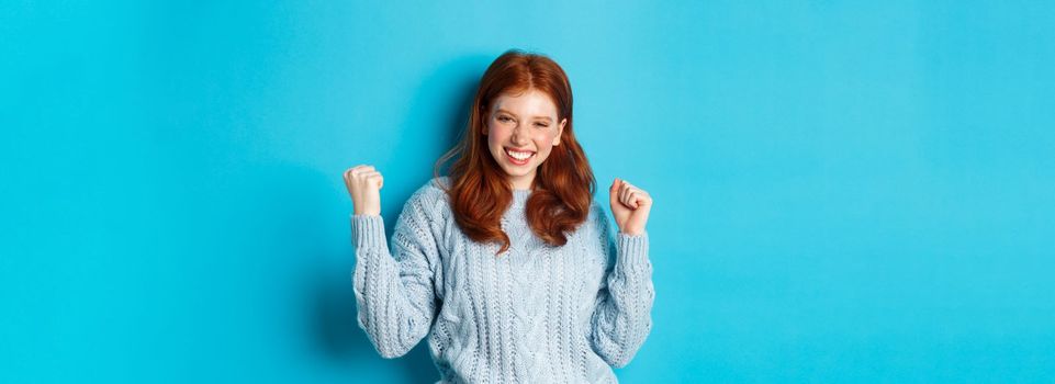 Satisfied redhead girl achieve goal and celebrating, making fist pump gesture and smiling delighted, triumphing of win, standing against blue background.