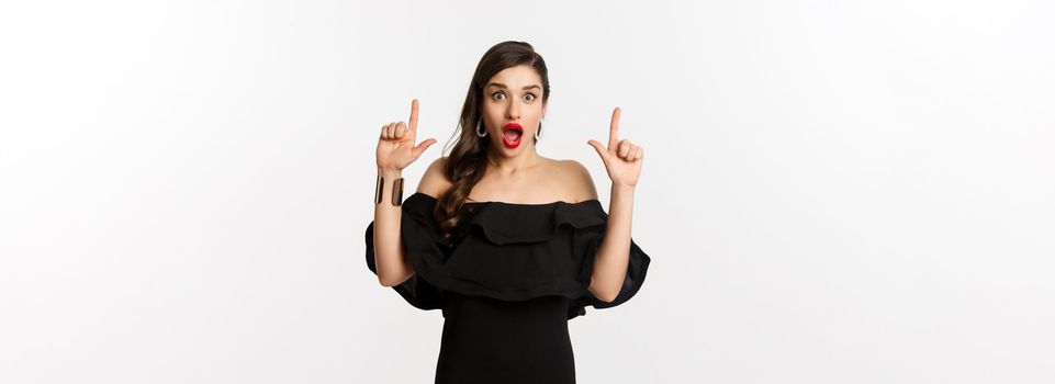 Fashion and beauty. Surprised woman in black dress pointing fingers up, showing banner, standing over white background.