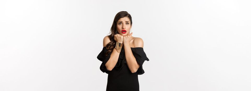 Fashion and beauty concept. Scared timid woman grimacing, looking concerned and worried at camera, standing over white background in black dress.
