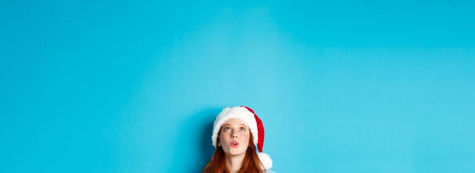 Winter holidays and Christmas eve concept. Head of pretty redhead girl in santa hat, appear from bottom and looking up at logo impressed, seeing promo offer, blue background.