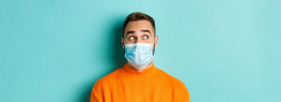 Covid-19, social distancing and quarantine concept. Close-up of young male model in medical mask looking left curious, staring at promo copy space, light blue background.