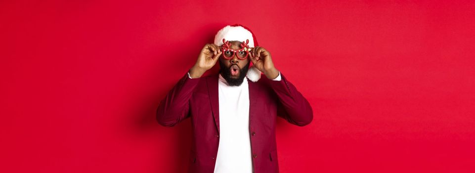 Funny Black man celebrating New Year, wearing party glasses and santa hat, having fun over red background.