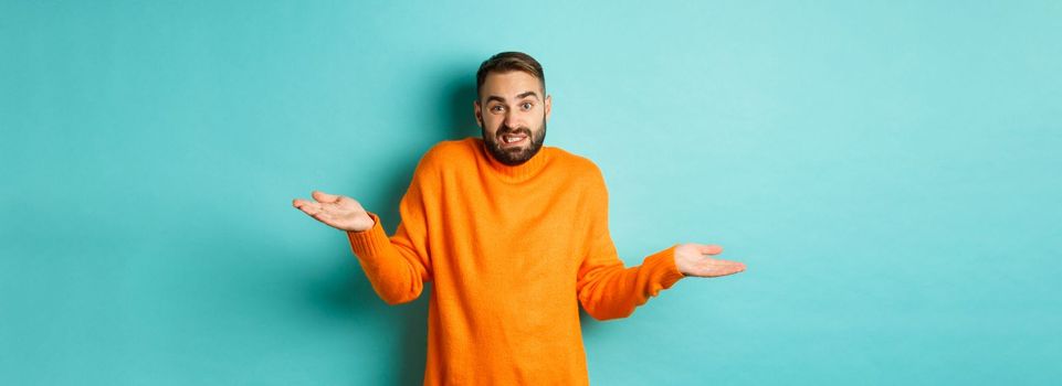 Confused caucasian man with beard, shrugging and looking clueless, standing indecisive against turquoise background.