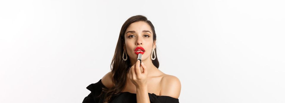 Close-up of beautiful woman applying red lipstick on lips, looking at camera like mirror, standing in black dress over white background.