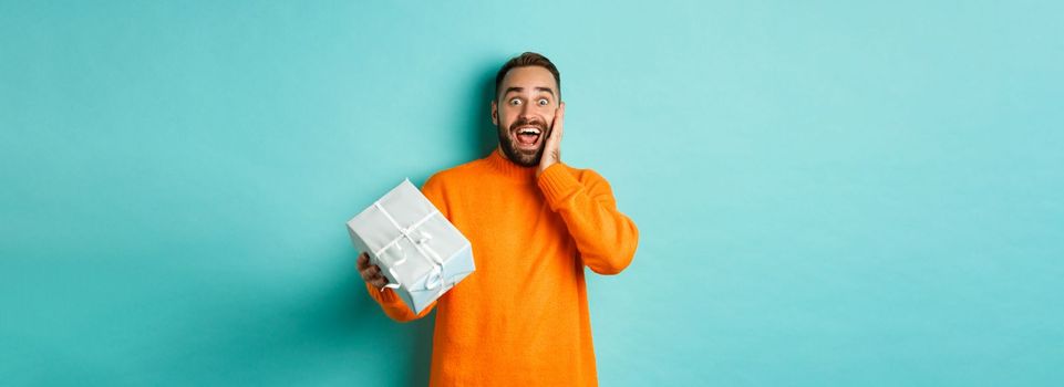 Holidays and celebration concept. Surprised man receiving gift, looking happy at present and smiling, standing over blue background.