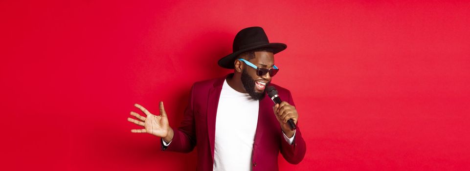 Passionate black male singer performing against red background, singing into microphone, wearing party outfit, standing over red background.