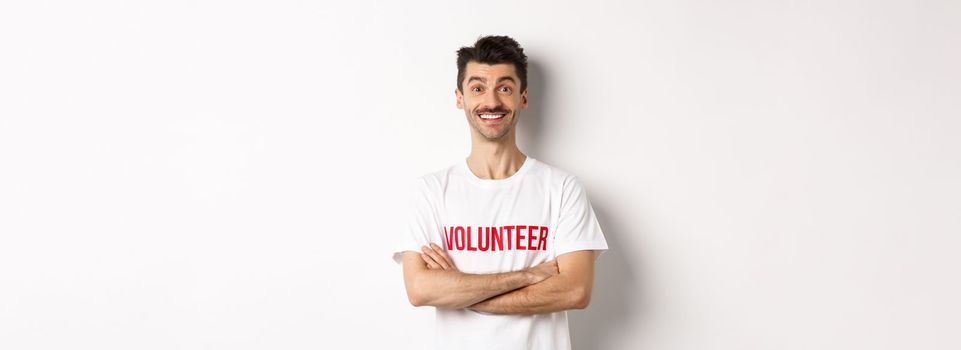 Happy young man in volunteer t-shirt ready to help, smiling at camera, cross arms on chest confident, white background.