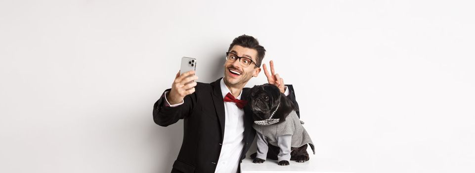 Cheerful dog owner in suit celebrating Christmas with dog, taking selfie on smartphone near cute black pug in costume, white background.