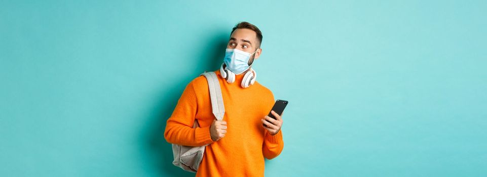 Young man in face mask using mobile phone, holding backpack, staring right amazed, standing against light blue background.