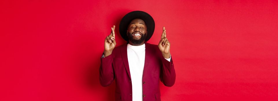 Hopeful african american man making wish, holding fingers crossed for good luck and smiling optimistic, standing against red party background.