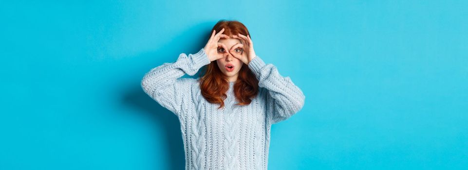 Funny redhead female model in sweater, staring at camera through fingers glasses, seeing something interesting, standing over blue background.