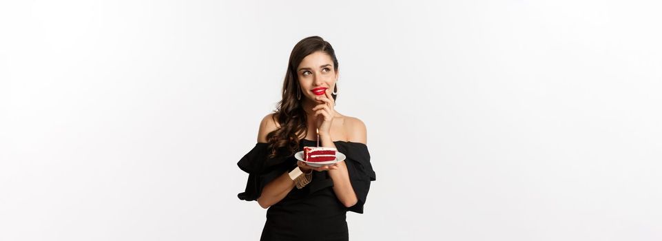 Celebration and party concept. Dreamy woman in black dress making wish, thinking and holding birthday cake with candle, standing over white background.