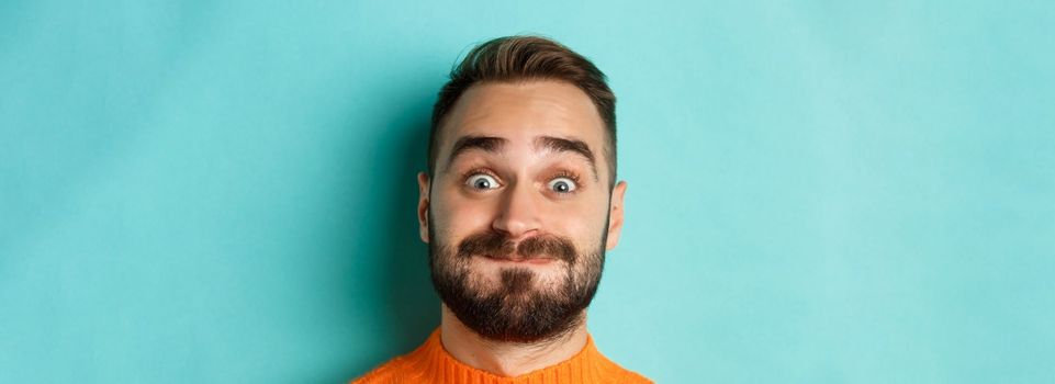 Headshot of funny bearded man showing faces and pouting, holding breath, standing in orange sweater against turquoise background.