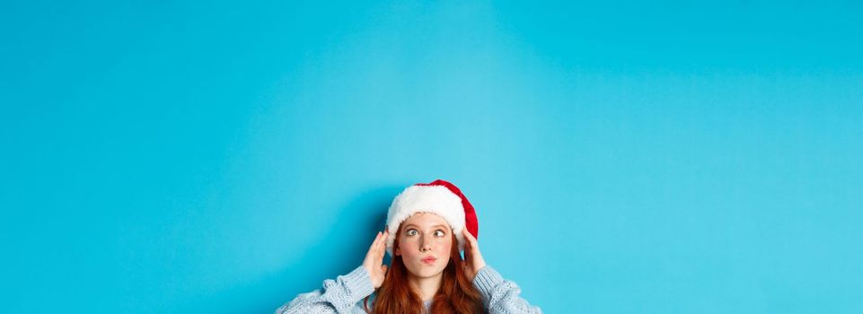 Winter holidays and Christmas eve concept. Head of funny redhead girl in santa hat, appear from bottom and squinting, making silly faces, standing near copy space on blue background.