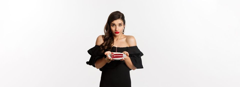 Party and celebration. Tender woman in black dress giving you birthday cake, asking to make wish on b-day candle, standing over white background.