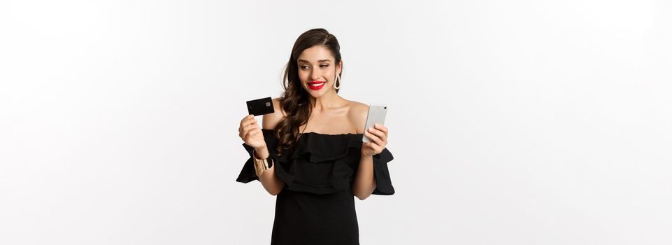 Fashion and shopping concept. Happy good-looking woman buying online, holding credit card and smartphone, white background.