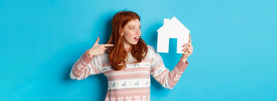 Real estate concept. Excited redhead female with red hair, pointing and looking at paper house model, showing apartment advertisement, standing over blue background.