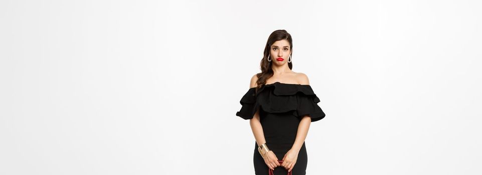 Beauty and fashion concept. Full length if silly young woman pouting and looking confused, holding purse, wearing heels and black dress, white background.