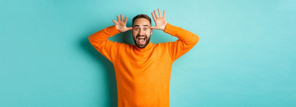 Image of handsome caucasian man making funny faces, mocking someone and smiling, standing against light blue background.