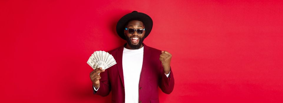 Cheerful african american guy in sunglasses, black hat and blazer, winning prize money, holding dollars and looking satisfied, standing over red background.
