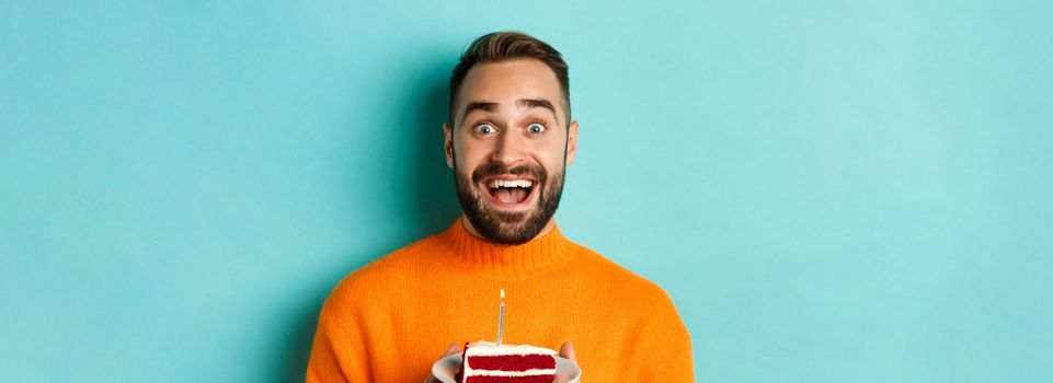 Close-up of happy adult man celebrating birthday, holding bday cake with candle and making wish, standing against turquoise background.