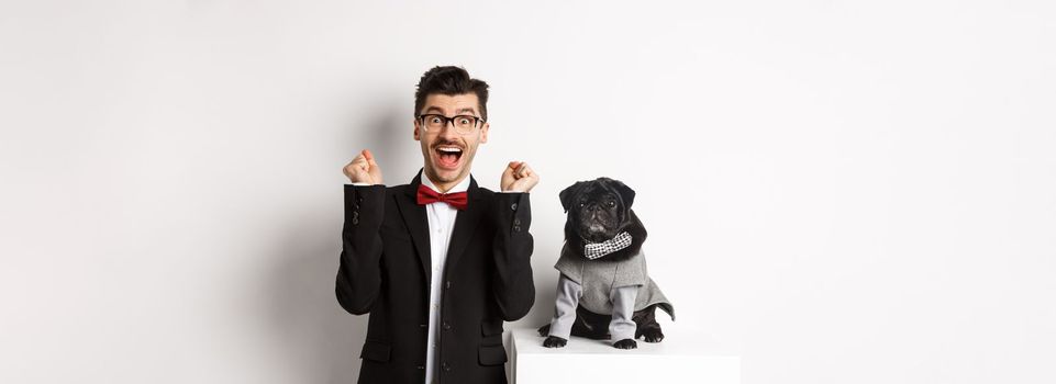 Animals, party and celebration concept. Happy young man in suit and puppy in pet cosume standing over white background, dog owner rejoicing and triumphing.