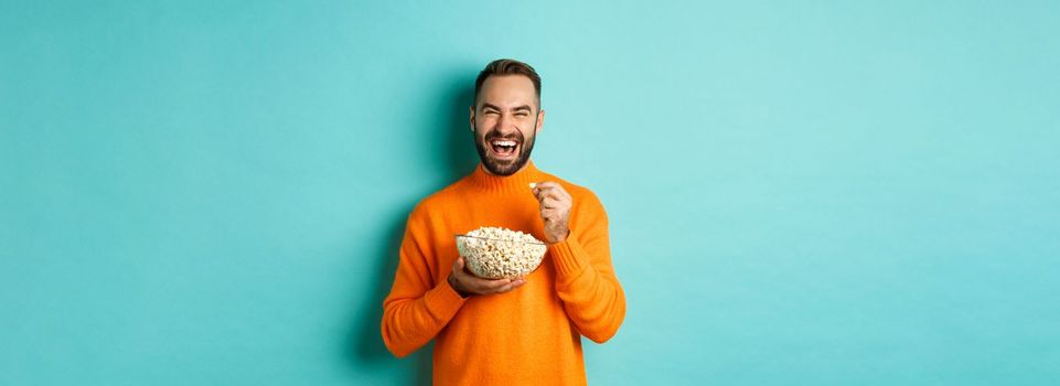 Excited young man watching interesting movie on tv screen, eating popcorn and looking amazed, blue background.