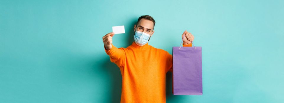 Covid-19, pandemic and lifestyle concept. Happy male customer in face mask showing credit card and purple shopping bag, standing over light blue background.