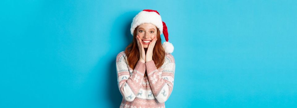 Winter and Christmas Eve concept. Happy redhead girl celebrating xmas, staring at camera amazed, smiling and touching cheeks, standing over blue background.