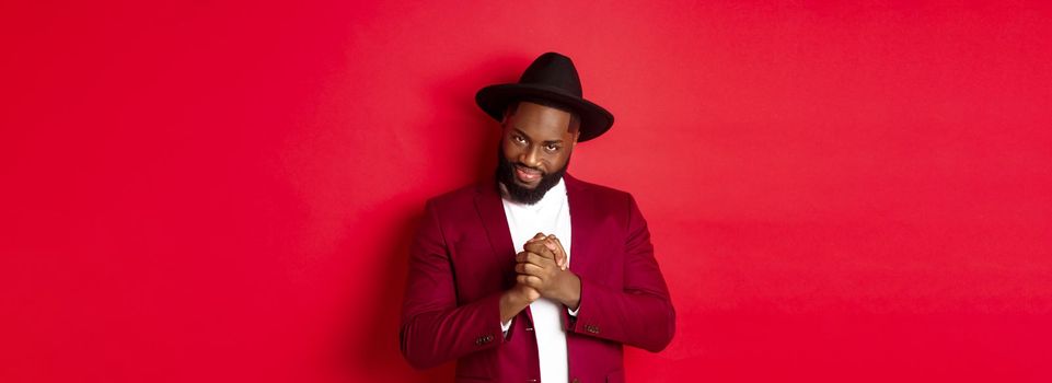 Cunning black man having an idea, smiling devious and rubbing hands, standing against red background and scheming.