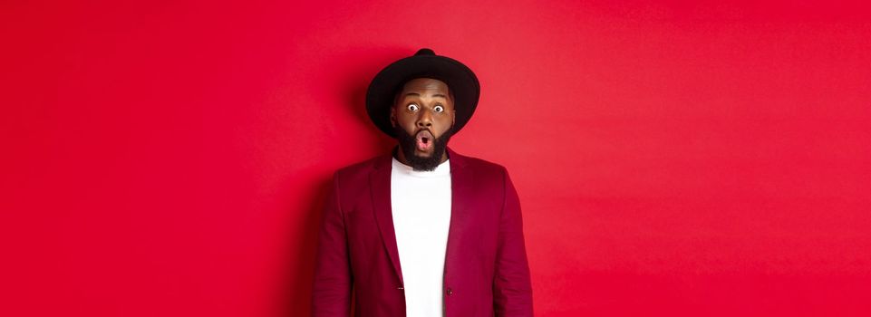 Fashion and party concept. Impressed Black man in classy outfit staring with complete disbelief at camera, gasping and looking surprised, standing over red background.