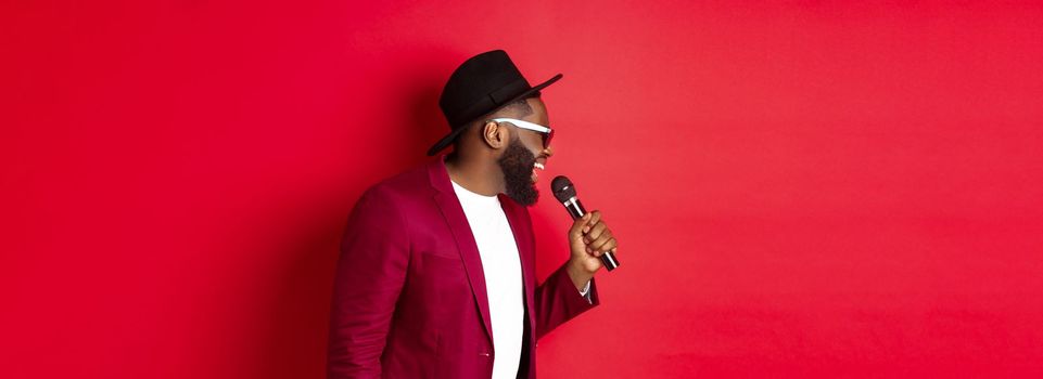 Passionate black male singer performing against red background, singing into microphone, wearing party outfit, standing over red background.