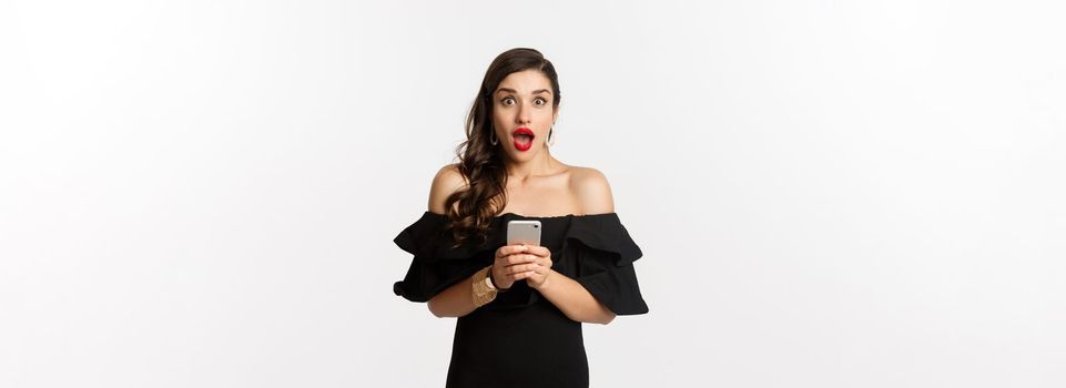 Happy young glamour woman in black dress, holding mobile phone and looking surprised, standing over white background.