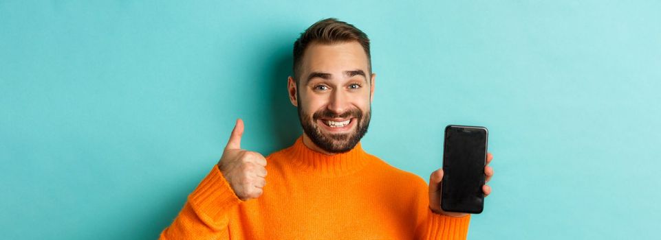 Close-up of attractive bearded man showing smartphone screen, thumbs up, recommending mobile app, standing satisfied over turquoise background.