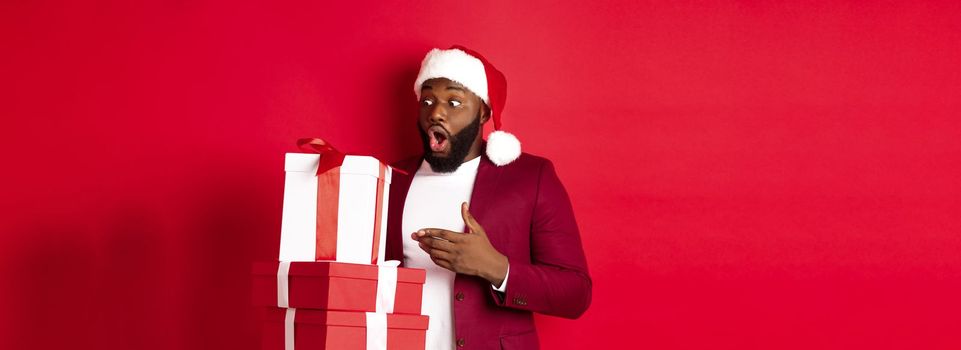 Christmas, New Year and shopping concept. Surprised Black man looking at xmas presents with amazement, wearing santa hat, standing with gifts against red background.